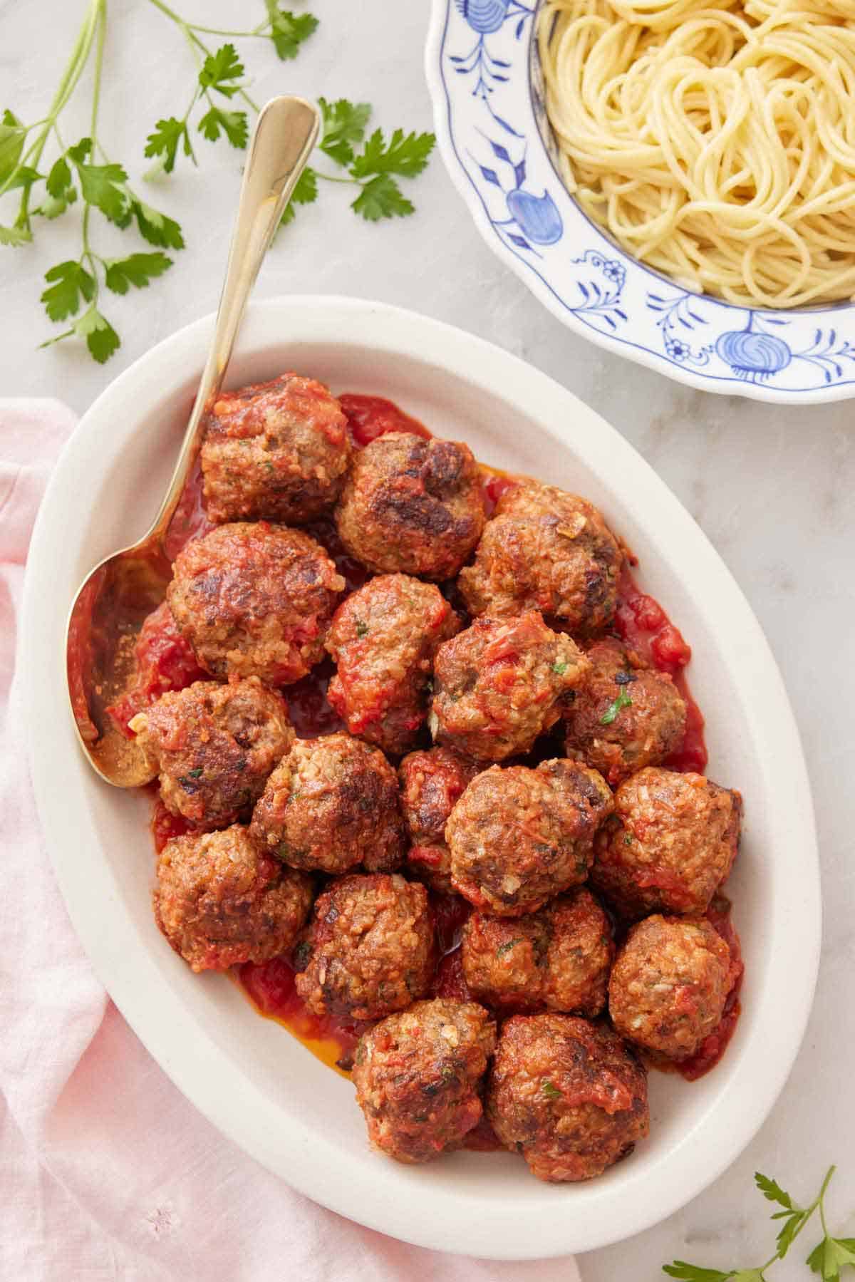 Overhead view of a white platter with meatballs tossed in sauce with a plate of spaghetti beside it.