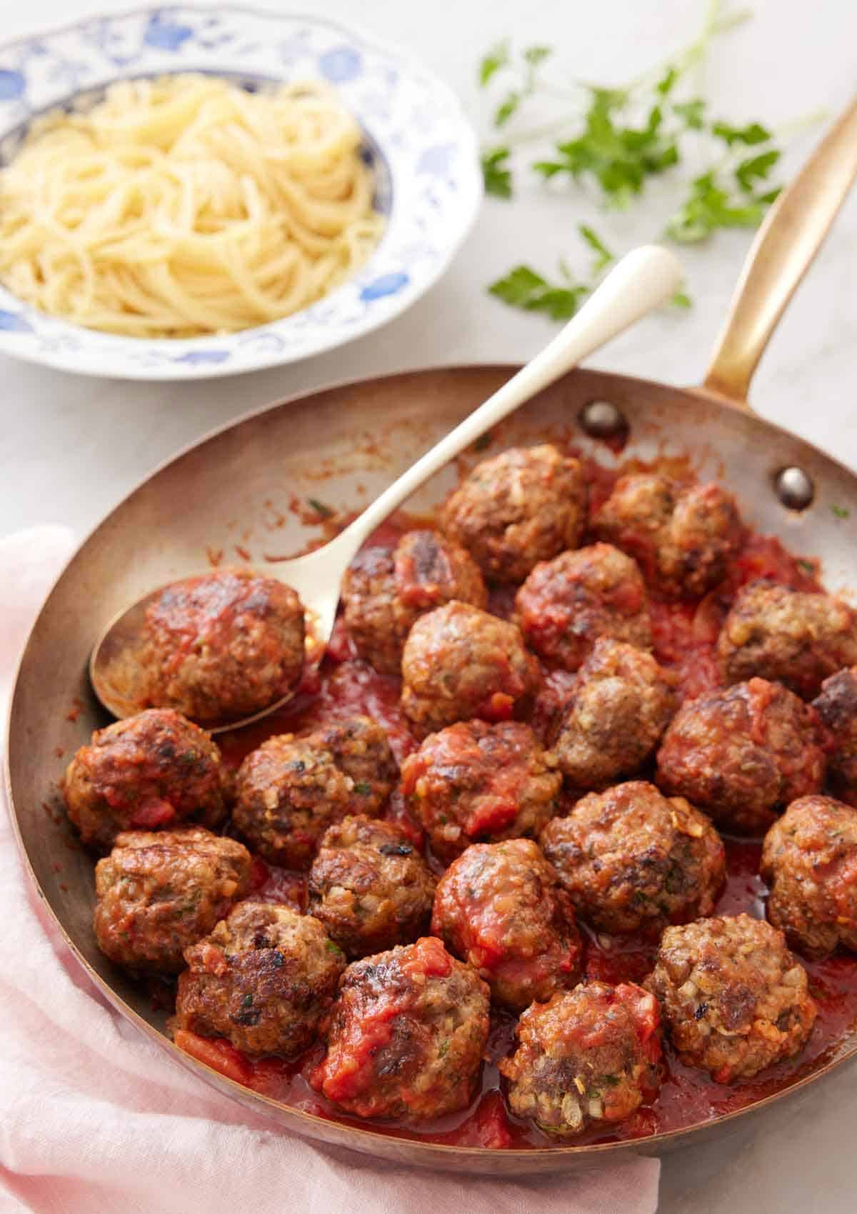 A skillet of meatballs with sauce and a spoon with some noodles in the background.