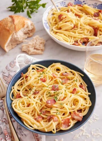 A bowl of pasta carbonara with torn bread, glass of wine, and another bowl of pasta in the back.