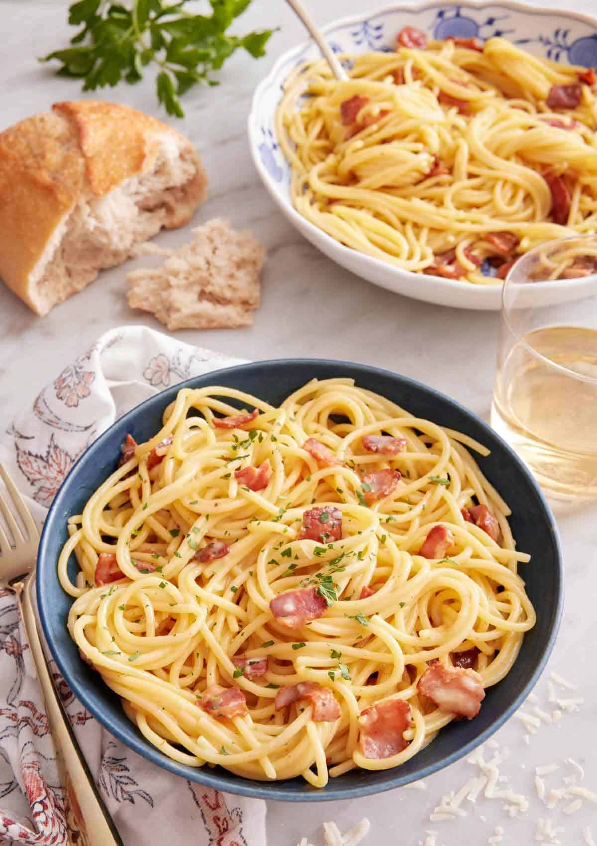 A bowl of pasta carbonara with torn bread, glass of wine, and another bowl of pasta in the back.