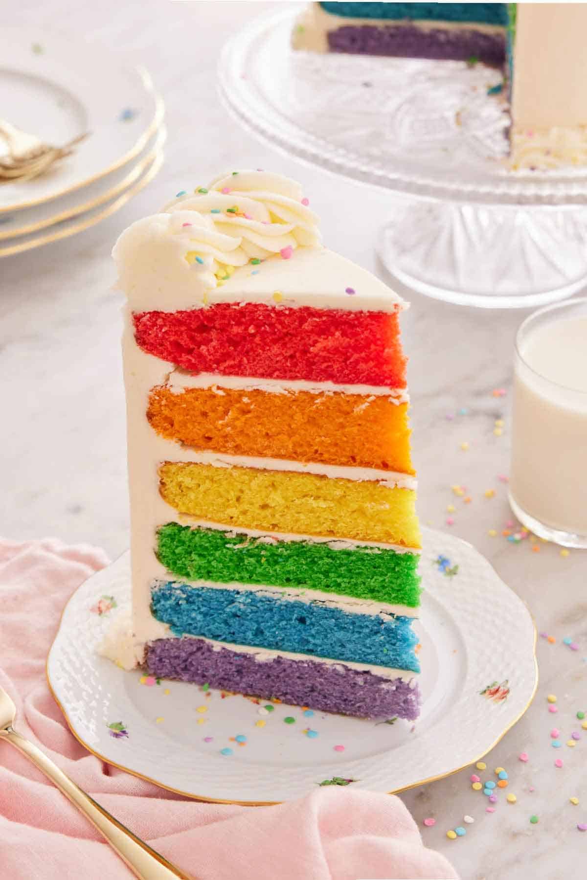 A slice of rainbow cake on a plate with rainbow sprinkles on top and scattered around.
