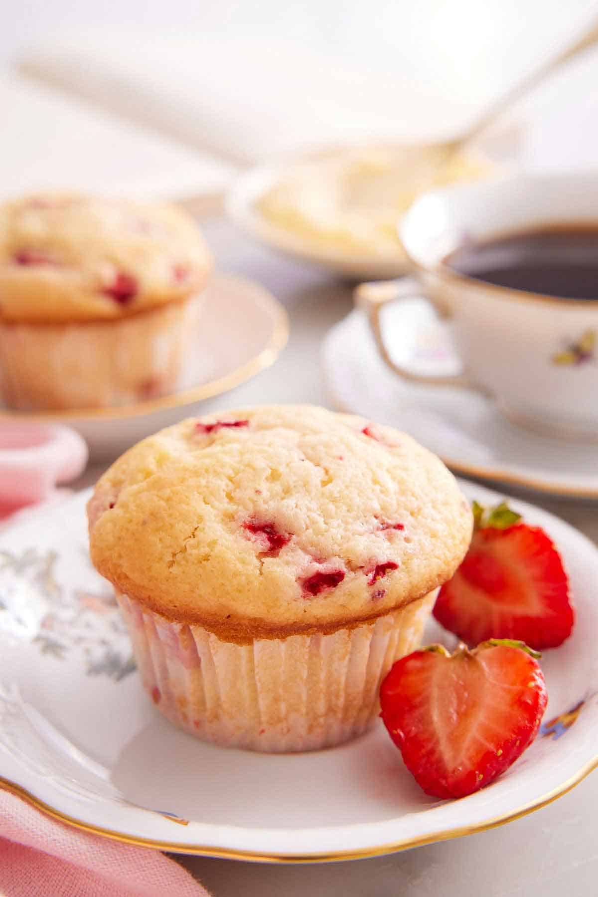 A plate with a strawberry muffin with a cut strawberry with a cup of coffee and second muffin in the background.