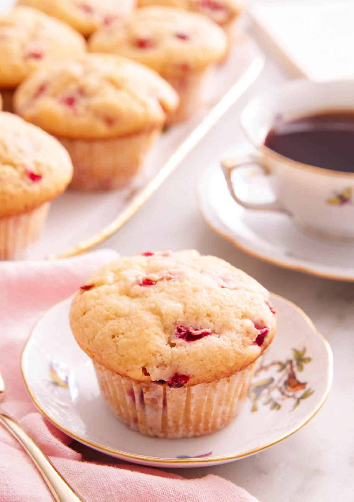 A strawberry muffin on a small plate with a cup of coffee and more muffins on a platter in the background.
