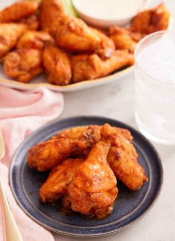 A plate with multiple air fryer chicken wings with a platter of more in the background with a glass of ice water.