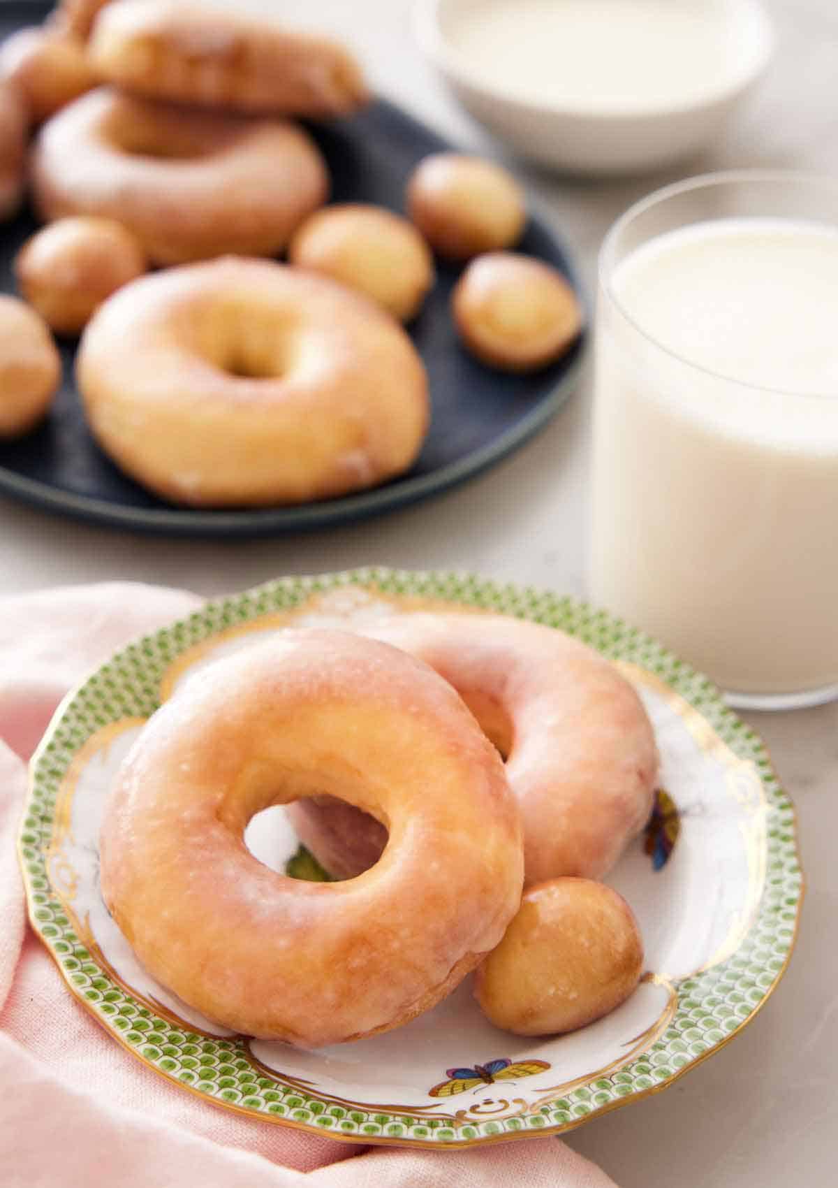 A plate with two air fryer donuts and a donut hole with a glass of milk behind it and a platter of more donuts.