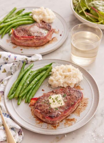 Two plates of bacon wrapped filet mignon with mashed potatoes and green beans with one plate in front of the other.