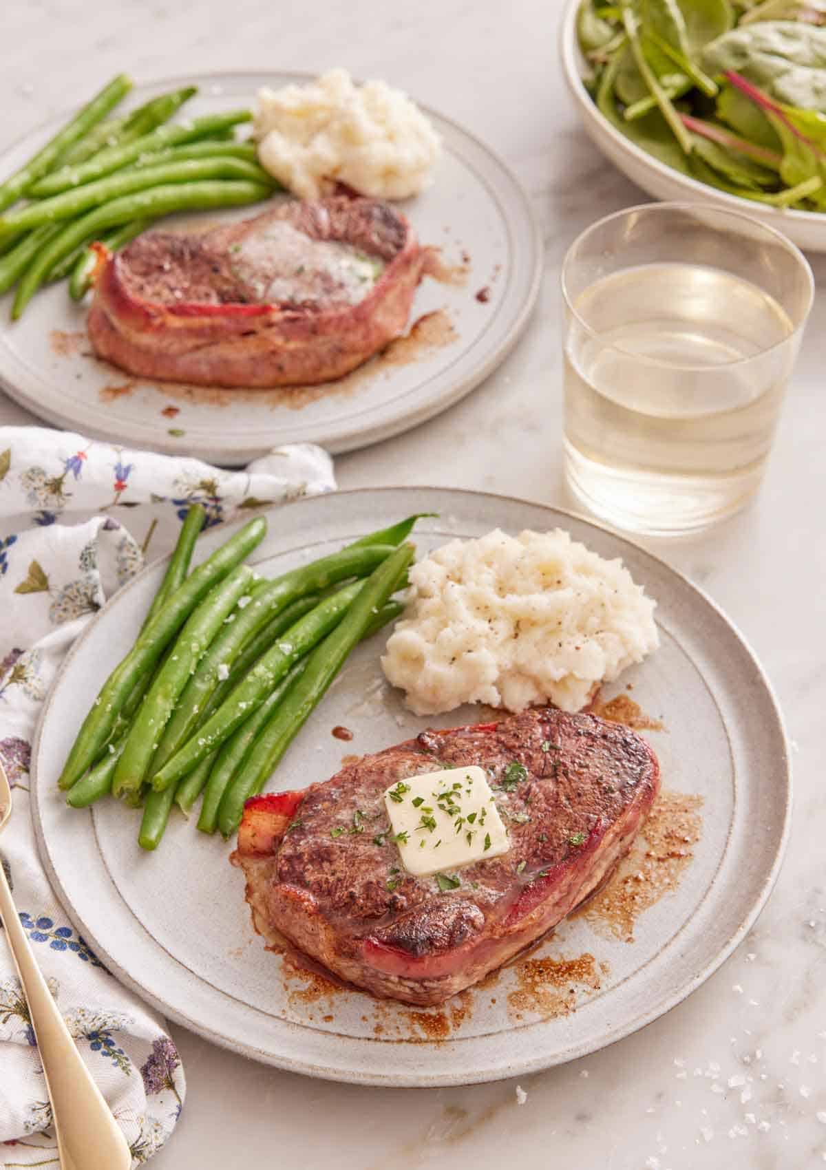 Two plates of bacon wrapped filet mignon with mashed potatoes and green beans with one plate in front of the other.