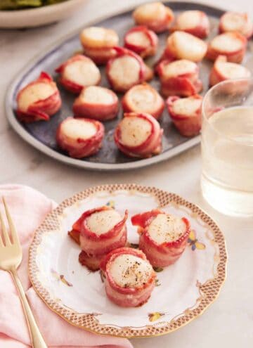 A plate with three bacon wrapped scallops with a platter in the back along with a glass of wine.