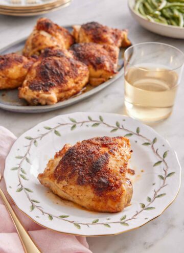 A plate with a baked chicken thigh with a glass of wine and additional chicken thighs in the background.