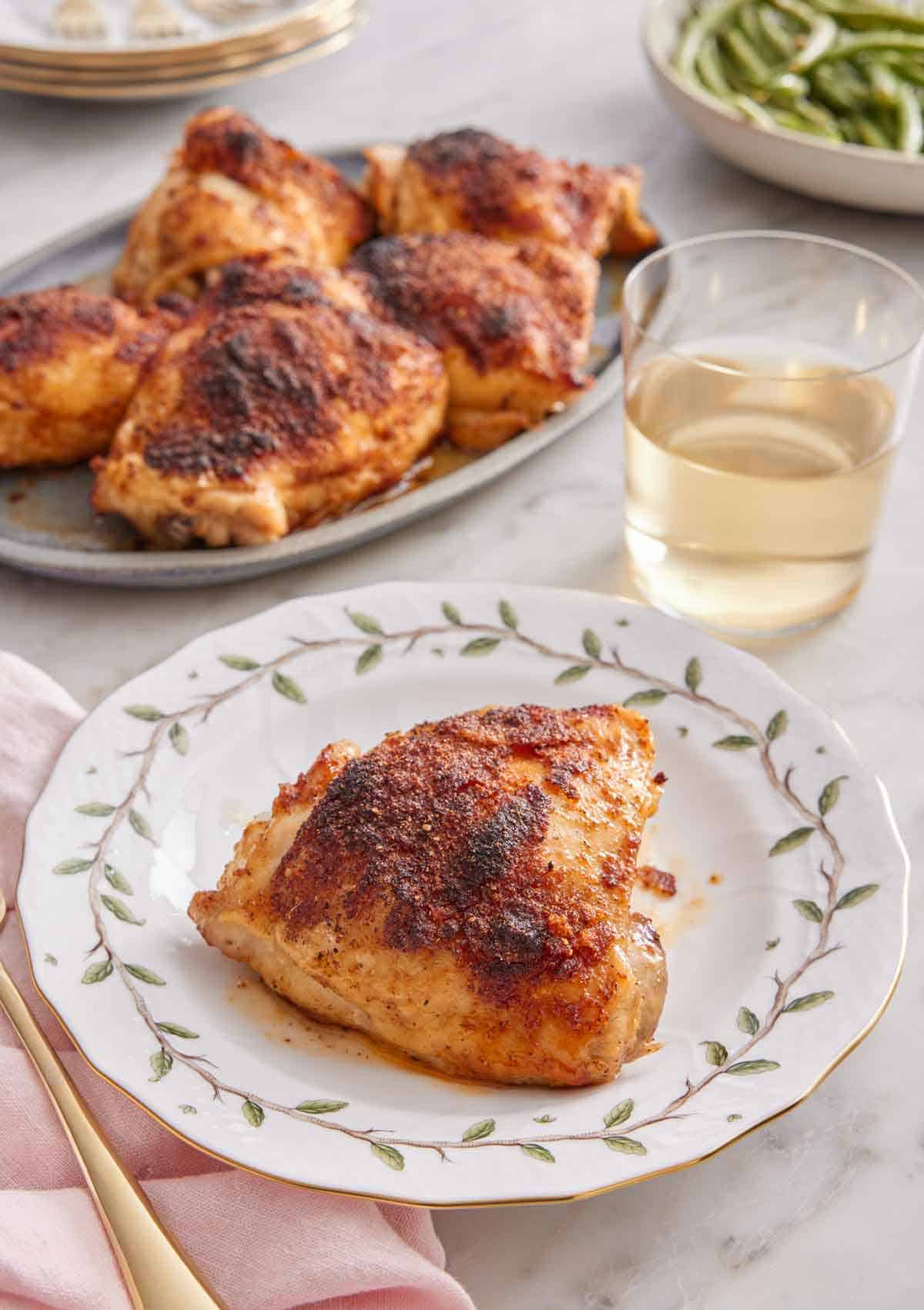 A plate with a baked chicken thigh with a glass of wine and additional chicken thighs in the background.