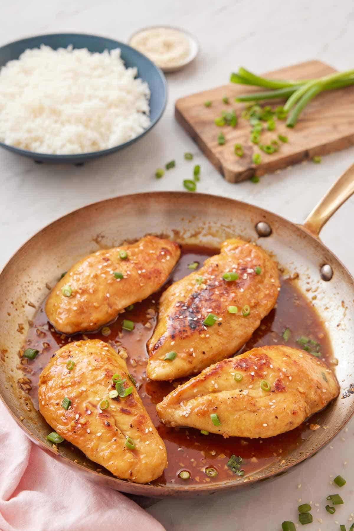 A skillet of honey garlic chicken with a cutting board with chopped green onions and a bowl of rice in the background.