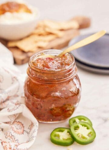 A mason jar of jalapeno jelly with a spoon inside and some sliced jalapeno in front.