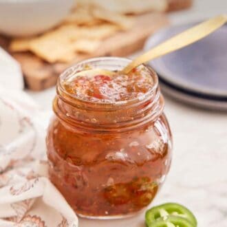 Pinterest graphic of a jar of jalapeno jelly with a spoon tucked in with three sliced of jalapeno peppers in front.