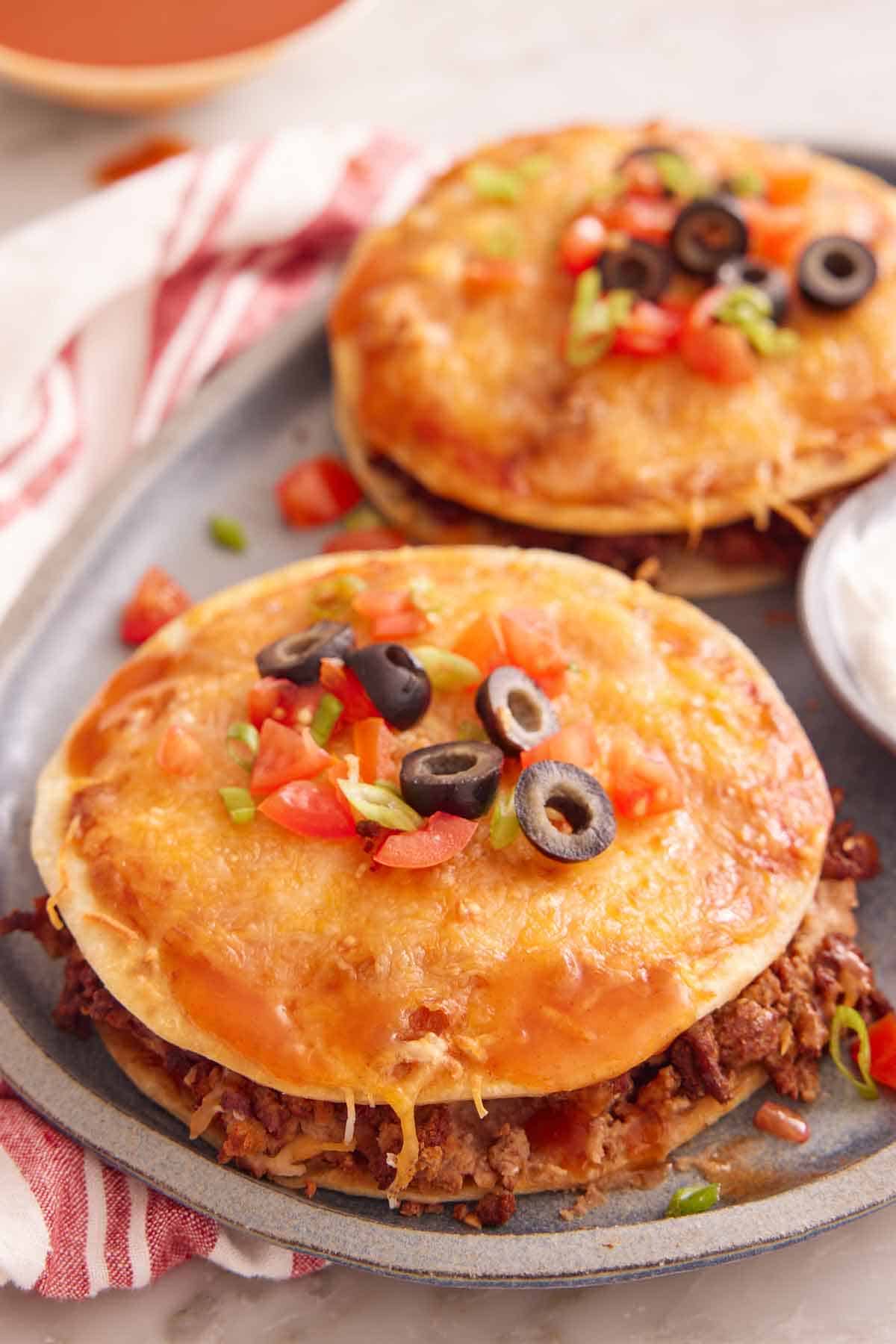 A Mexican pizza with diced tomatoes, olives, and green onions on top with a second one in the background.