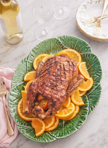 Overhead view of a green platter with a roasted duck overtop sliced oranges. Wine glasses and wine bottle in the background.