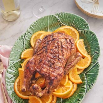 Pinterest graphic of an overhead view of a green platter with a roasted duck overtop sliced oranges with wine glasses and wine bottle in the background.