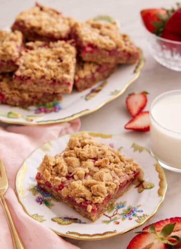 A plate with a slice of strawberry rhubarb bar with a glass of milk and a platter of bars in the background.