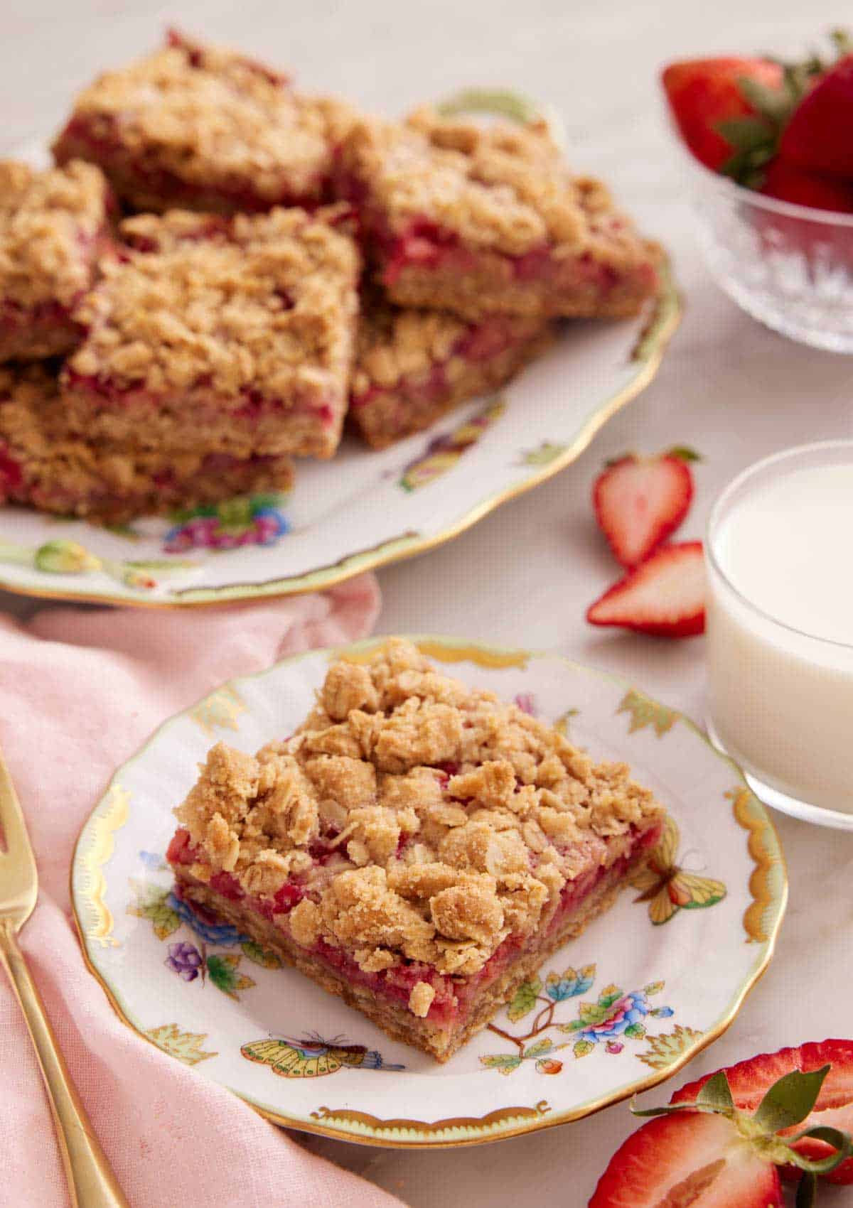A plate with a slice of strawberry rhubarb bar with a glass of milk and a platter of bars in the background.