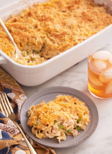 A plate with a serving of tuna casserole with a baking dish full in the back and an iced drink on the side.