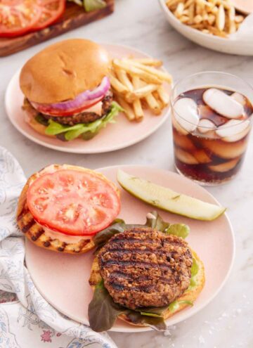 A plate with an opened veggie burger with a drink on the side with another veggie burger in the background with fries.