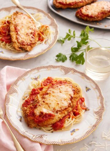 Two plates of air fryer chicken parmesan over spaghetti noodles with a glass of wine and parsley in between the plates.