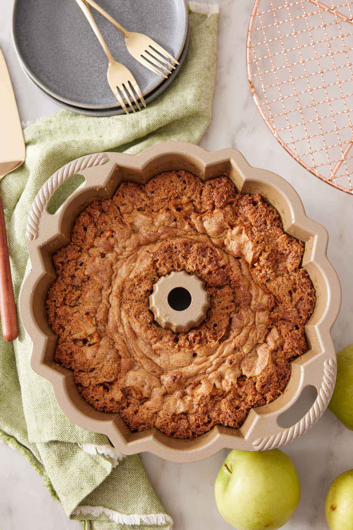 Overhead view of apple bundt cake in a bundt pan. A cooling rack, stack of plates and forks, and apples off to the sides.