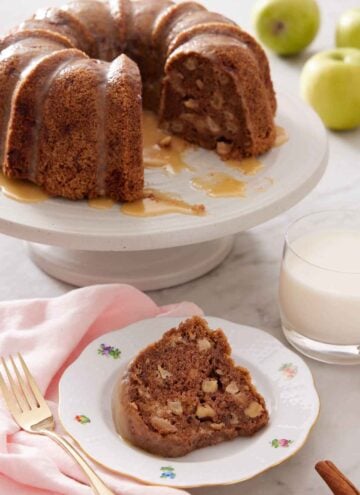 A plate with a slice of apple bundt cake with a cake stand in the background holding the rest of the cake.