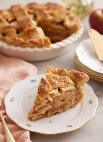 A slice of apple pie on a plate in front of the rest of the pie.