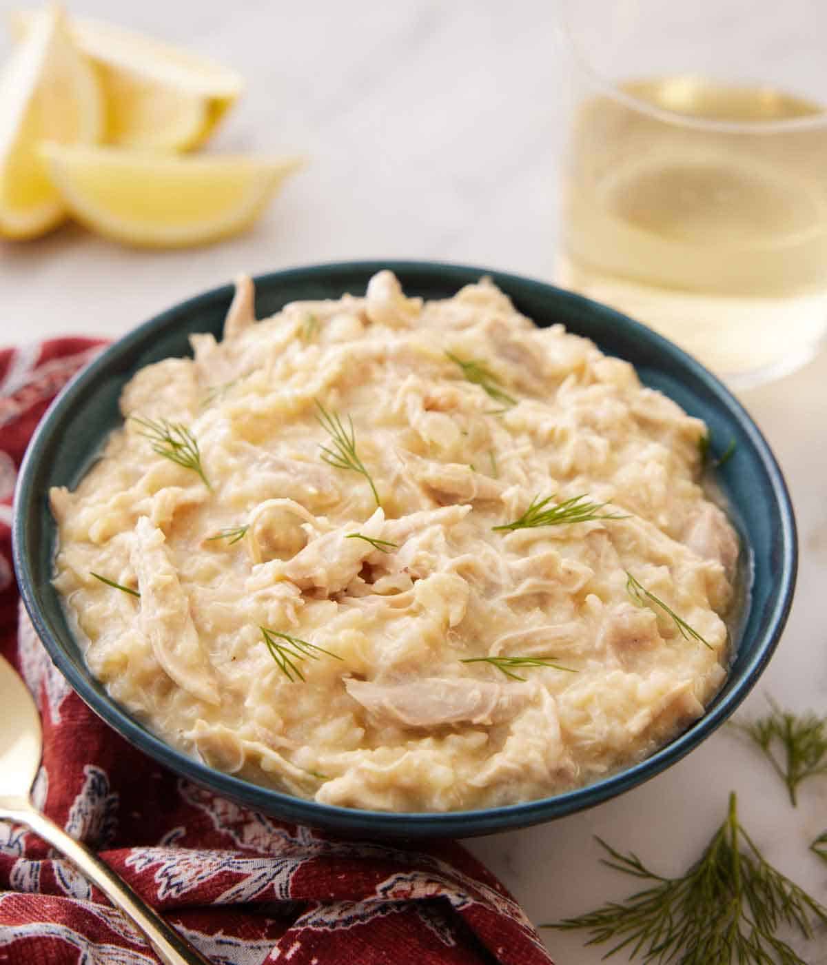 A blue bowl of avgolemono with a drink in the background along with lemon wedges.