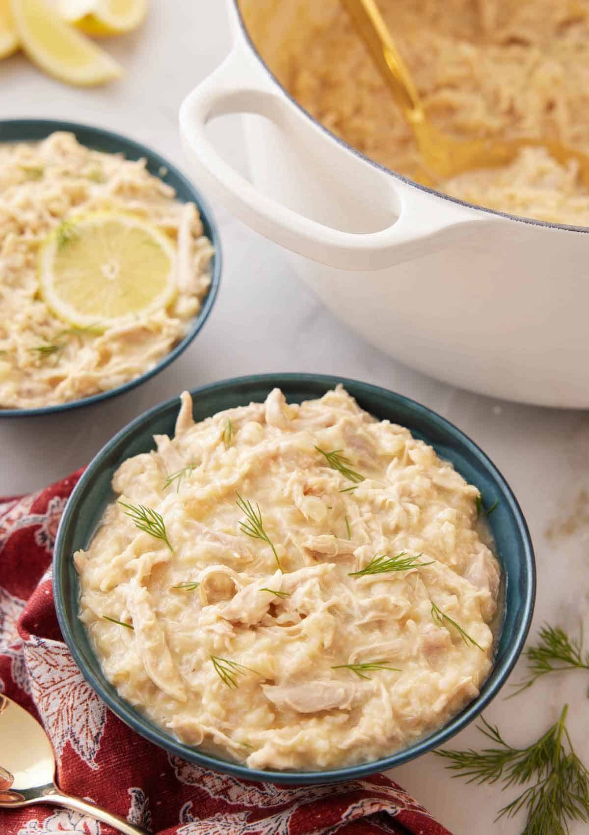 A bowl of avgolemono with a large pot and second bowl of avgolemono in the background.