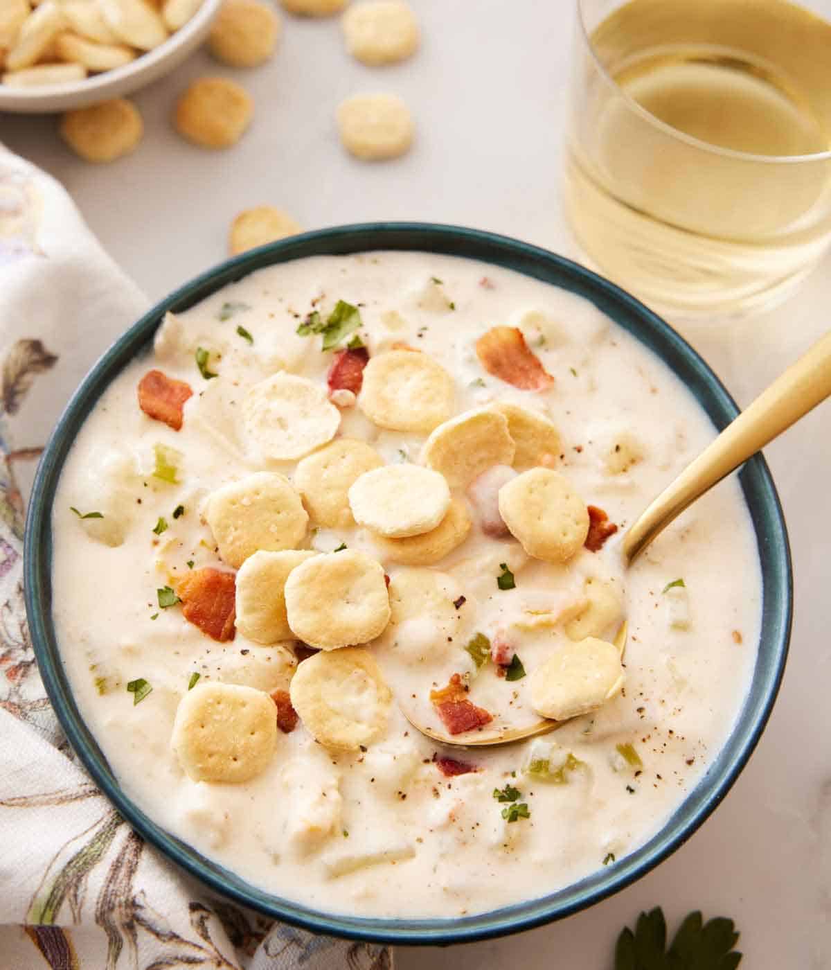 A bowl of clam chowder with crackers on top with a glass of wine in the back.