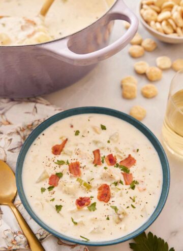 A bowl of clam chowder with diced bacon on top with crackers and a purple pot in the background.