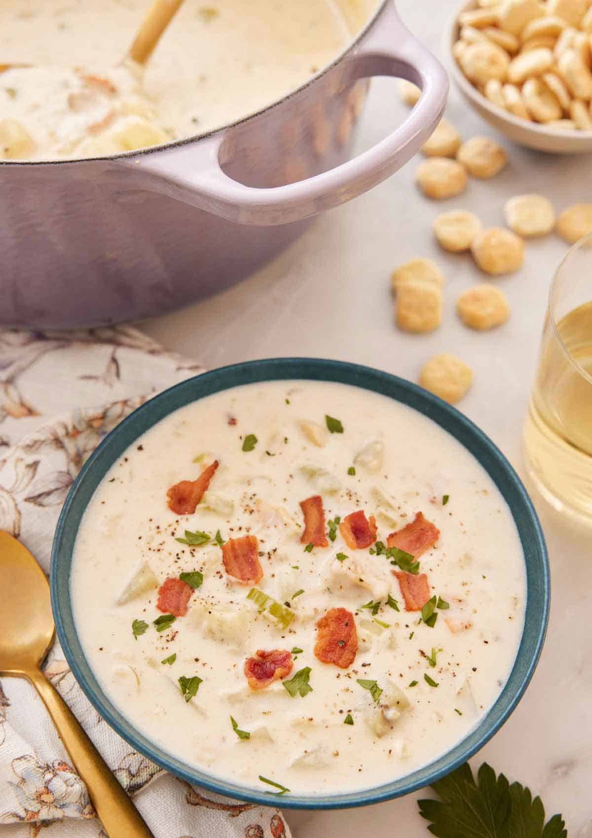 A bowl of clam chowder with diced bacon on top with crackers and a purple pot in the background.