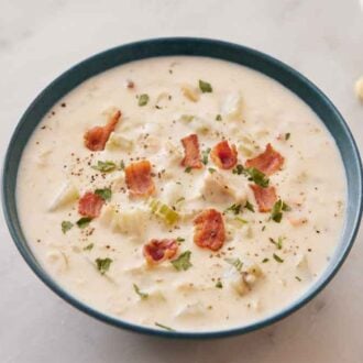 A blue bowl of clam chowder with bacon on top and crackers on the side.