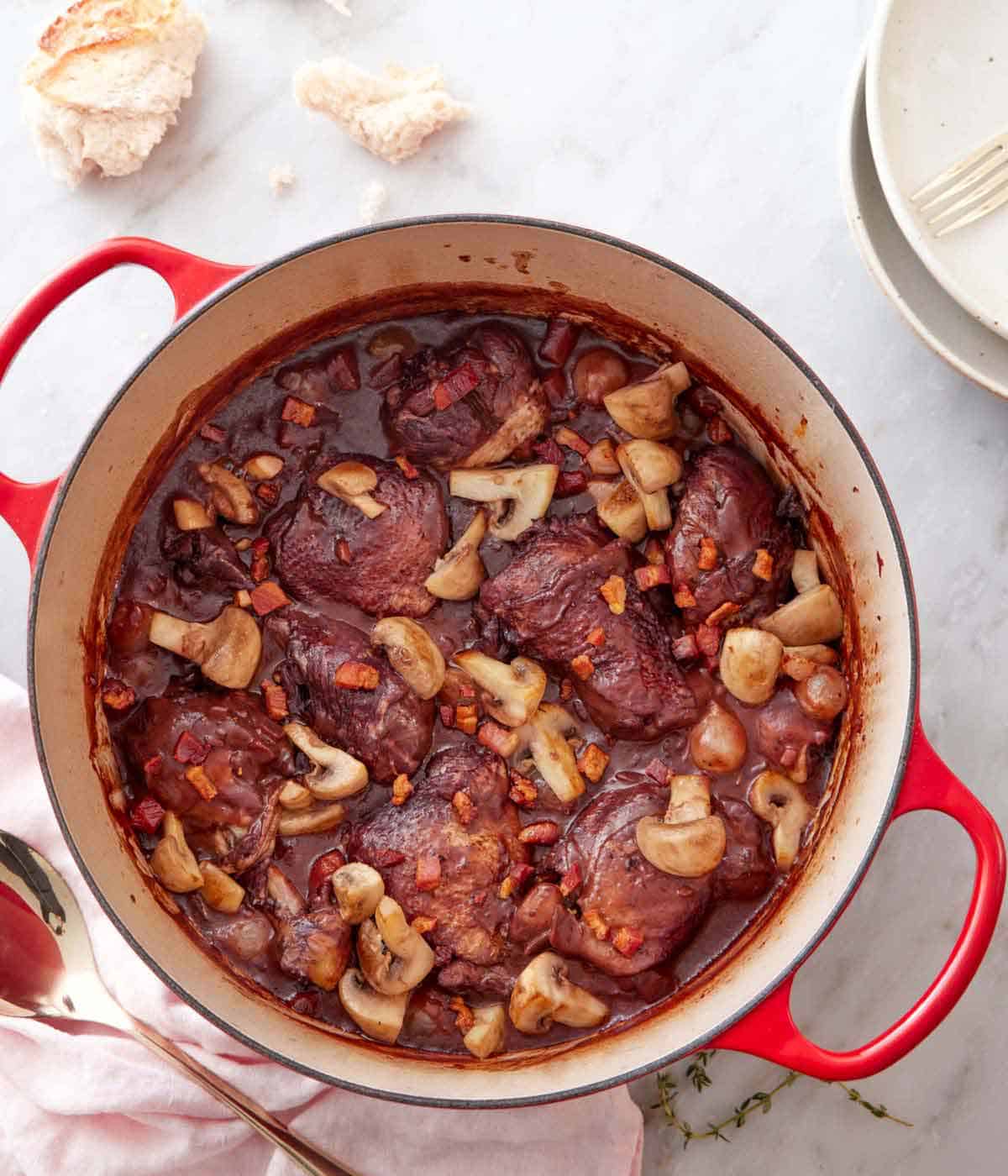 Overhead view of a pot of coq au vin with some torn bread and plates off to the side.