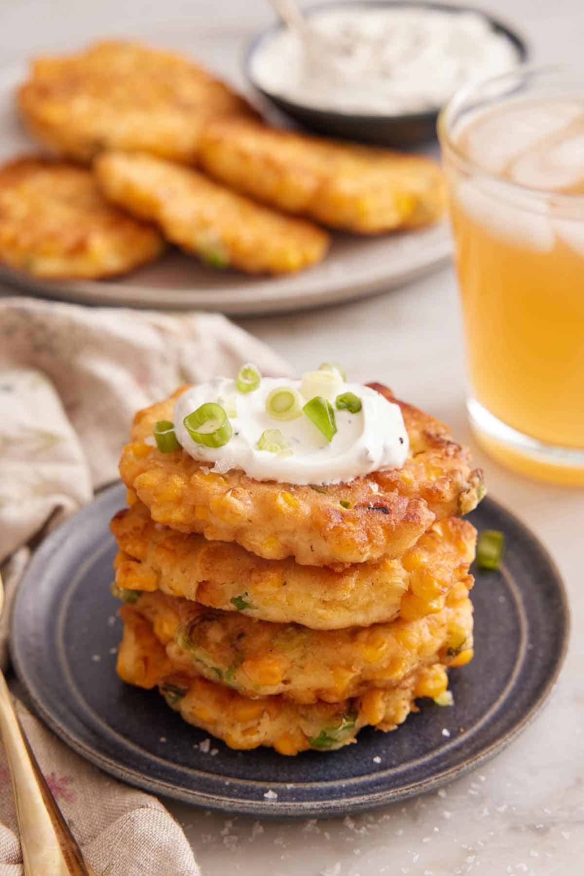 A plate with a stack of four corn fritters with sour cream on top and garnished with green onions. An iced drink in the back along with a platter of additional corn fritters.