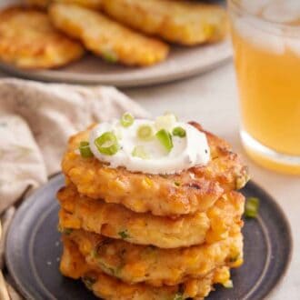 Pinterest graphic of a plate with a stack of four corn fritters with sour cream on top and garnished with green onions. An iced drink in the back along with a platter of additional corn fritters.
