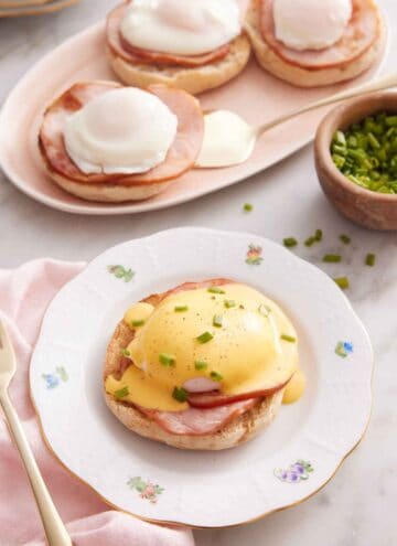 A plate with eggs benedict with hollandaise sauce over top with some chives. More eggs benedicts in the background.