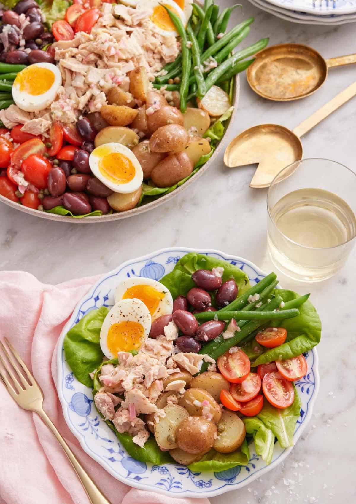A plate of niçoise salad with a glass of wine in the back with a platter of more niçoise salad.