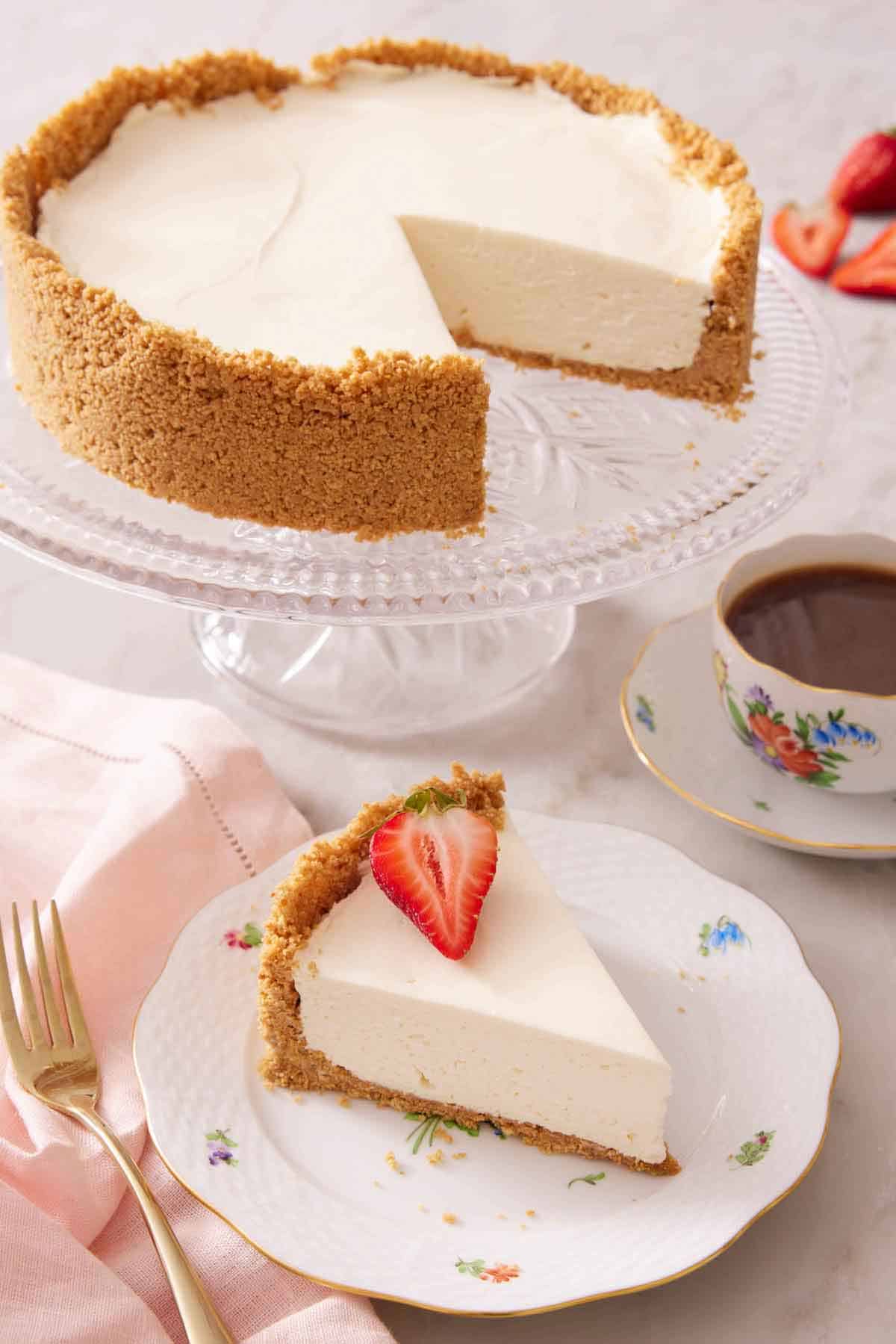 A plate with a slice of no bake cheesecake with a piece of strawberry on top with a cake stand with the rest of the cake.