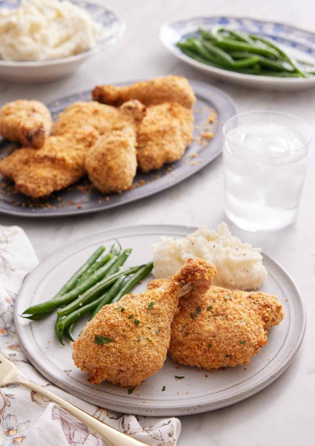 A plate of oven fried chicken with green beans and mashed potatoes. A glass of water and platters of food in the background.