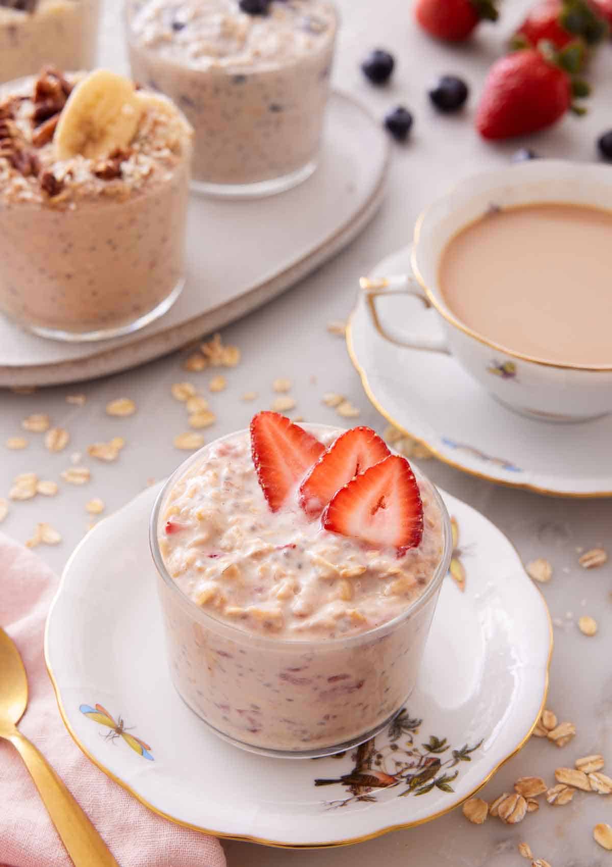 A plate with a glass of overnight oats topped with strawberries with a cup of coffee and more overnight oats in the background.