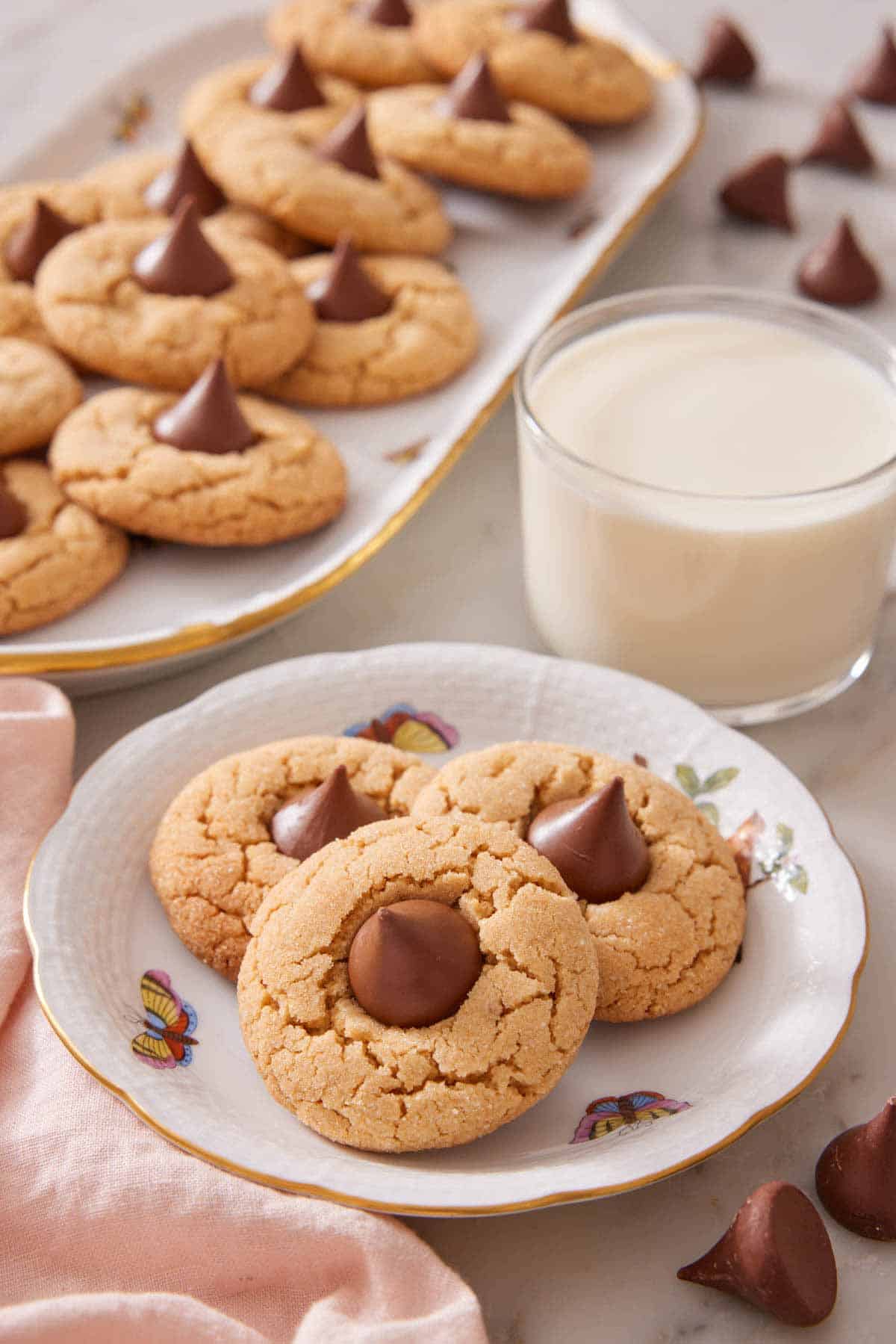 A plate with three peanut butter blossoms with a glass of milk and a platter of more peanut butter blossoms in the background.