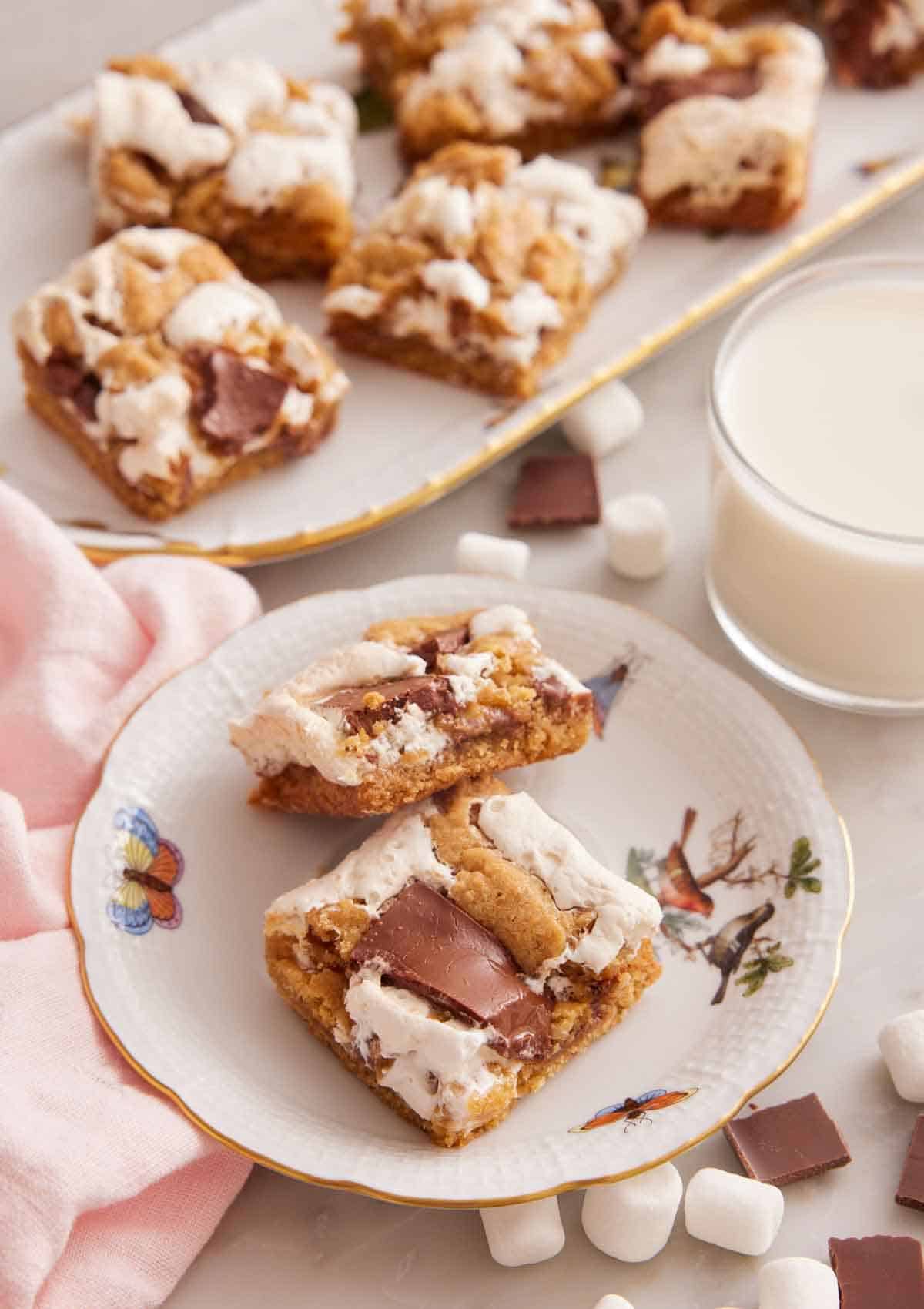 A plate with two pieces of s'mores bars with a cup of milk and platter of bars in the background.