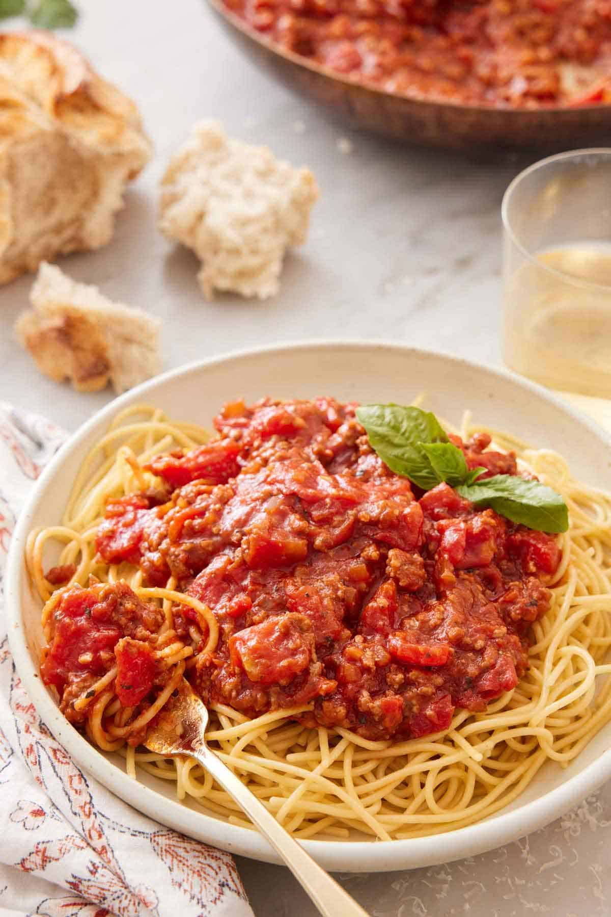 A bowl of noodles with spaghetti sauce on top and a fork tucked in. A drink and torn bread in the background.