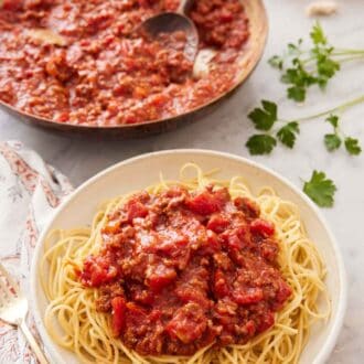 A bowl of noodles with spaghetti sauce on top with a skillet full of sauce in the background.