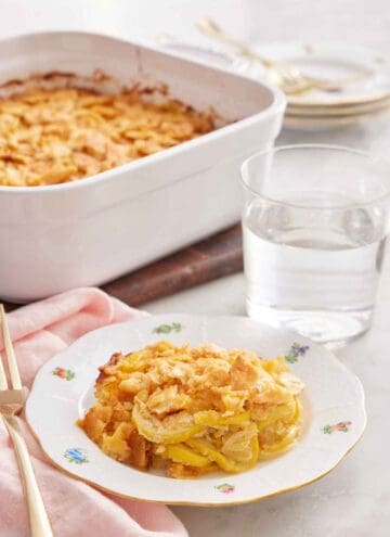 A plate with a serving of squash casserole with a glass of water and baking dish in the background.