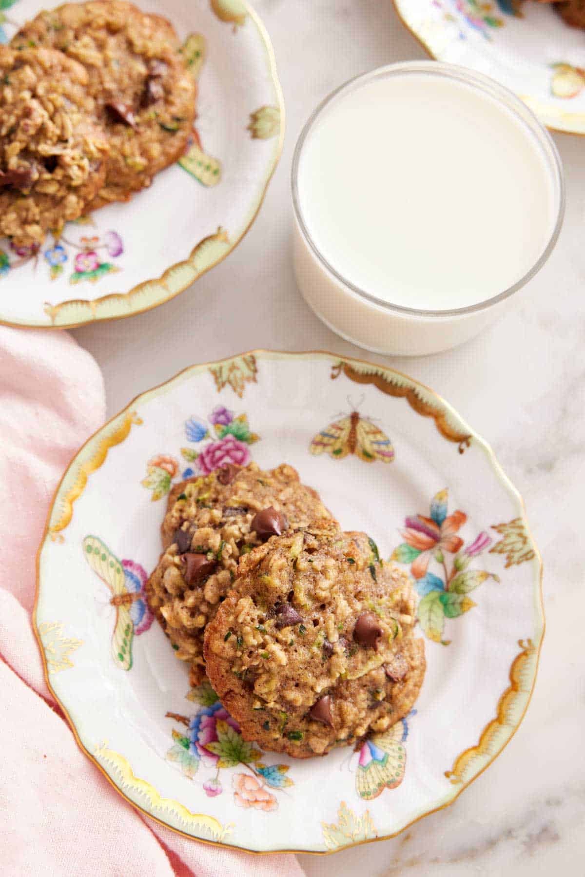 Overhead view of a plate with two zucchini cookies with a glass of milk and another plate of cookies.