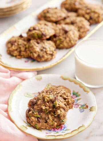 A plate with two zucchini cookies with a glass of milk and platter of more cookies in the background.