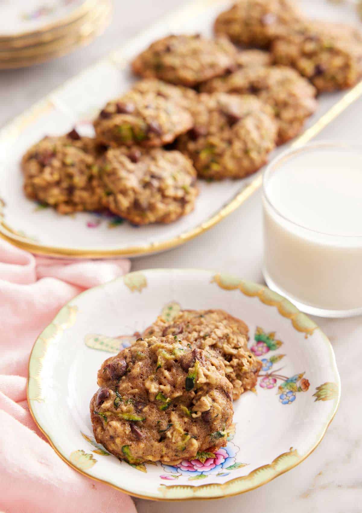A plate with two zucchini cookies with a glass of milk and platter of more cookies in the background.